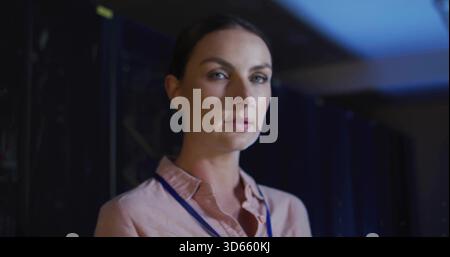 Standing woman wearing pink shirt and lanyard monitoring racks LEDs cables in blue-lit data center Stock Photo
