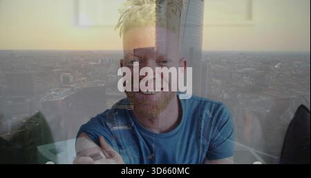 Smiling man with blond dreads and beard wearing blue tee leaning in flat with skyline reflection Stock Photo