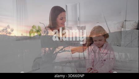Smiling mother and daughter sitting on bed in home bedroom, with pink bathrobe and sunset overlay Stock Photo