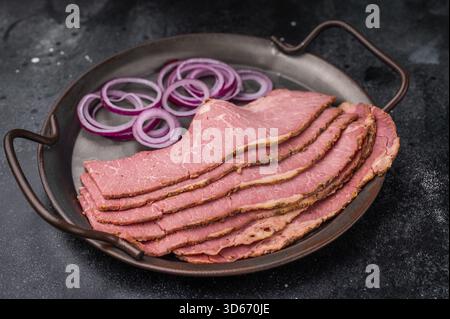 Thinly sliced pastrami on a metal plate with red onion rings and pickled cucumbers, placed on a textured dark stone background. Smoked deli beef ready Stock Photo