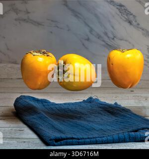 three fresh persimmons floating above blue cloth on wooden surface Stock Photo