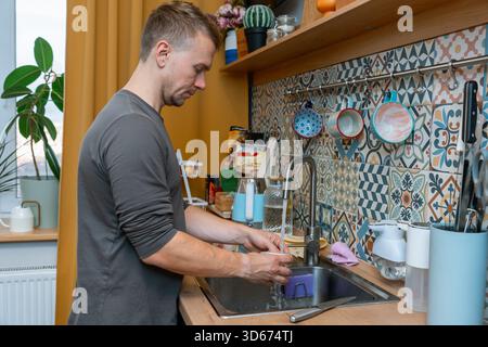 A young man washes dishes in the kitchen sink Stock Photo