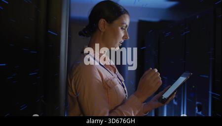 Holding tablet, tech inspecting LED-lit racks at data center, wearing pink shirt and lanyard Stock Photo
