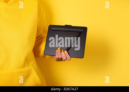 Person holding a digital graphic tablet and stylus, matching yellow hoodie and nail polish, isolated on vibrant yellow background. Stock Photo