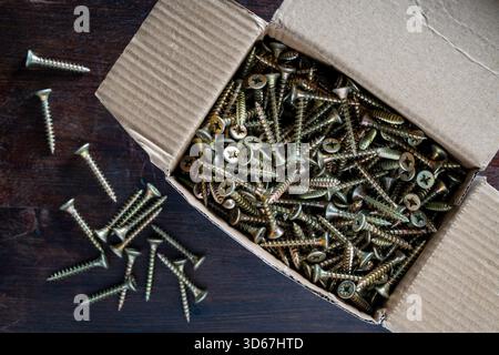 Many golden metal self tapping screws for construction work in a cardboard box on wooden background , close up, top view, copy space. New fasteners se Stock Photo
