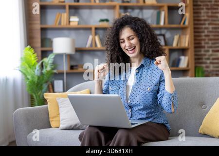 Woman feeling excited and happy, celebrating a win or achievement while working remotely on a laptop, sitting comfortably on a couch in a cozy home environment Stock Photo