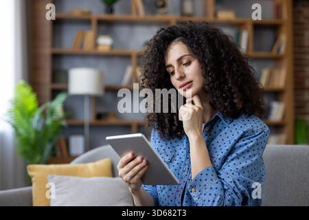 Woman with curly hair sitting on a couch, holding a digital tablet, intently engaging with content, using technology for online learning, entertainment, or remote work in a comfortable living room Stock Photo