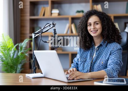 Young woman with curly hair hosting a podcast in her modern home office, smiling while using a laptop and professional microphone for remote work and online communication Stock Photo