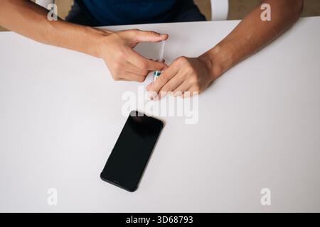 Top view of skilled repairman using syringe needle to repair broken smartphone in professional workshop sitting at white table. Stock Photo