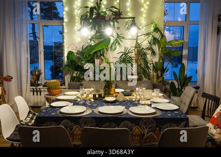 A festive table set for guests for Christmas and New Year, in the living room, plates, bottles, glasses, potted plants, fairy lights Stock Photo