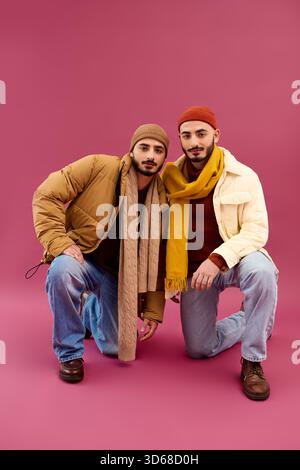 Two handsome twin brothers dressed in stylish attire kneel against a bright pink backdrop. Stock Photo
