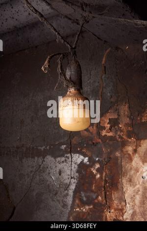 close-up of dirty light bulb fixture, yellow plastic diffuser caked in grime and soot hanging from dark ceiling, neglect and industrial decay concept Stock Photo