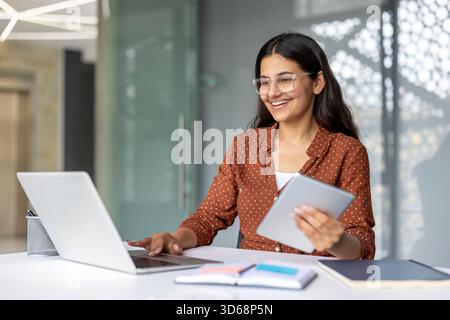 Young smiling indian woman multitasking, using a laptop and digital tablet for work, showing happiness and productivity in a modern office environment Stock Photo