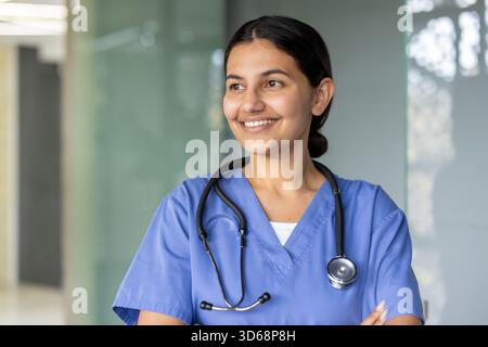 Young woman working as a professional nurse or doctor, wearing blue scrubs and a stethoscope, smiling while confidently looking away in a hospital setting Stock Photo