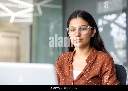 Focused indian woman wearing clear frame glasses and a brown polka dot shirt, concentrating on a laptop screen while working in a contemporary office environment Stock Photo