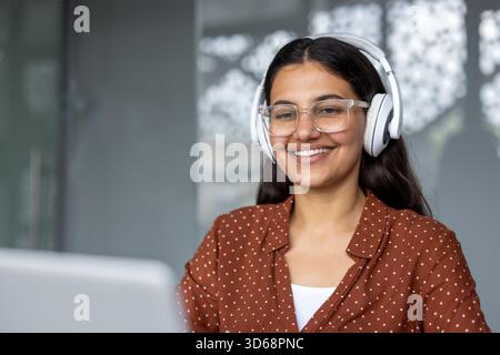 Young indian woman wearing white headphones and clear eyeglasses, smiling while looking at a laptop, engaging in remote work, online education, or a video call Stock Photo