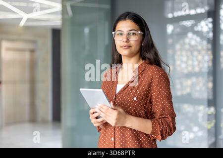 Young indian businesswoman wearing glasses and a polka dot shirt, standing confidently in a modern office, holding a digital tablet and looking at the viewer Stock Photo