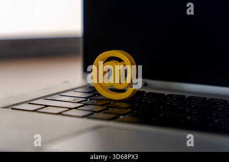 A yellow at sign placed on a laptop keyboard with stacked books in the background. Email, digital communication, internet, online services, and modern Stock Photo