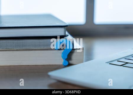 A small blue question mark placed between stacked books and a laptop. Inquiry, research, education, technical support, online study, confusion concept Stock Photo
