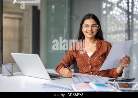 Young indian woman accountant smiling, holding documents and writing notes while working on a laptop at her desk in a modern office, representing finance, business, and success Stock Photo