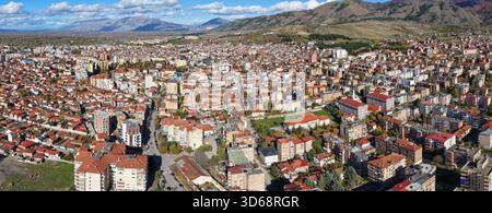 Wide aerial panorama of Korçë showing dense urban streets, mixed buildings, and dramatic mountain ranges under bright autumn skies. Stock Photo