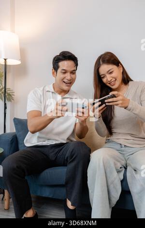 A cheerful Asian couple sitting together on a sofa, playing mobile games and laughing happily in a cozy living room. They enjoy their leisure time and Stock Photo