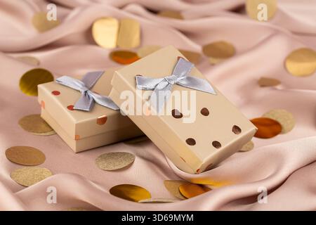 Two beige gift boxes with golden dots and gray ribbons on the pink silk background. Round confetti on the pink silk background. Stock Photo