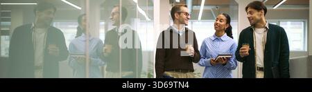 Three young adult colleagues including Caucasian man, Black woman, and Caucasian man walking in modern office hallway holding coffee cups and notebooks, engaging in conversation Stock Photo