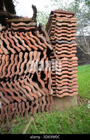 The photo shows a tall, uneven stack of terracotta roof tiles arranged in dense layers beside a tree, with green grass in the foreground and branches Stock Photo