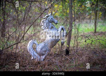 The photo shows a weathered silver horse statue standing on its hind legs in a patch of dry grass and twigs, surrounded by thin tree branches and gree Stock Photo