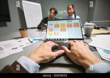 Caucasian young adult man working on laptop with project management software while multiethnic young adult colleagues discussing business strategy in modern office meeting room Stock Photo