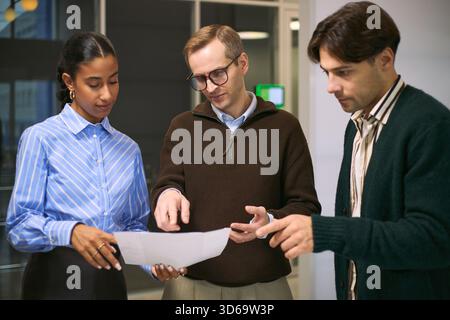 Multiethnic group of young adult business professionals discussing project details while reviewing documents in modern office setting, collaborating and sharing ideas together Stock Photo