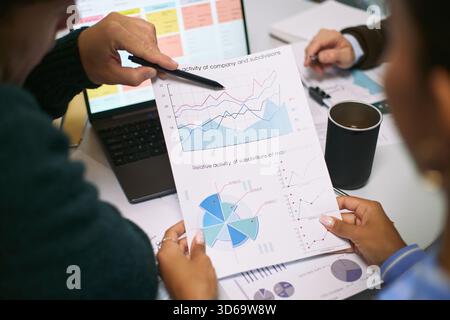 Caucasian man and Black woman analyzing financial charts and graphs during business meeting, discussing data trends and performance metrics with laptop and documents on desk Stock Photo