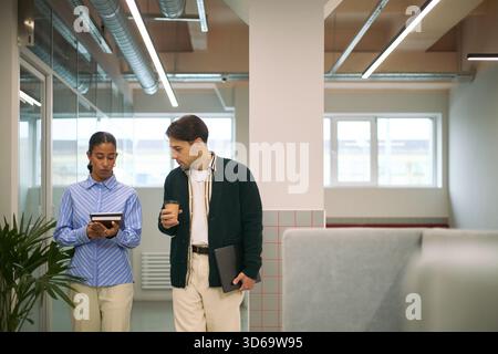 Young adult Black woman and young adult Caucasian man walking in modern office, woman holding digital tablet and man holding coffee cup and laptop, discussing business project Stock Photo