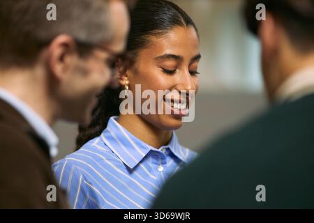 Young adult Black woman smiling while interacting with two colleagues during business meeting, standing in office environment, engaging in professional conversation, teamwork concept Stock Photo