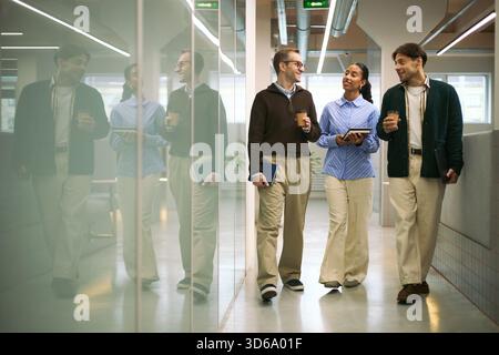 Three young adult colleagues, two Caucasian men and one Black woman, walking through modern office hallway holding coffee cups and digital tablet, engaging in conversation Stock Photo