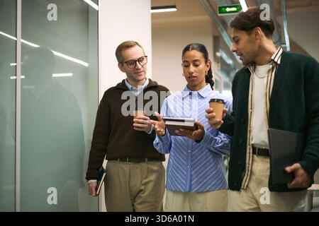 Multiethnic group of young adult professionals walking in office hallway discussing business strategy holding coffee cups and digital tablet collaborating on corporate project Stock Photo