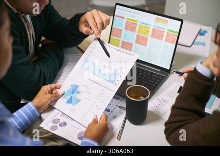 Three young adult men discussing financial charts and business analytics at desk, analyzing printed graphs and laptop screen with data, collaborating on project in office setting Stock Photo