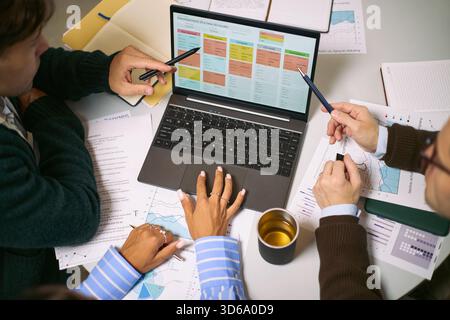 Diverse group of young adult and middle aged professionals collaborating around laptop, analyzing business charts and discussing project strategy in modern office setting Stock Photo