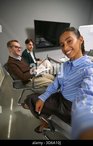 Young adult Black woman taking selfie with smiling young adult Caucasian man and young adult Caucasian man sitting in modern office during business meeting Stock Photo