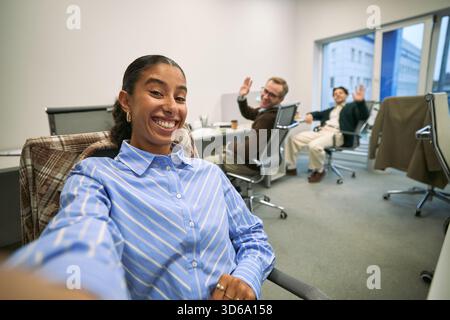 Portrait of young adult Hispanic woman smiling and taking selfie in modern office while two Caucasian men, one middle aged and one young adult, sitting at desks waving in background Stock Photo
