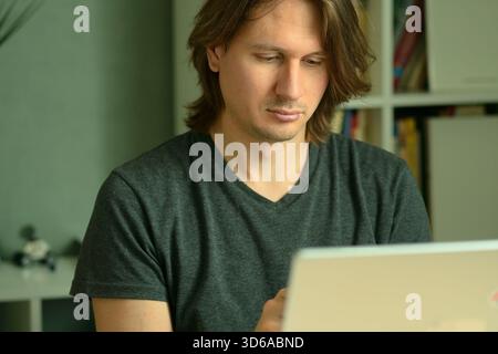 A young man with long hairstyle working on his laptop at home office desk. Focused look at the computer screen. Remote work and telecommuting concept. Stock Photo