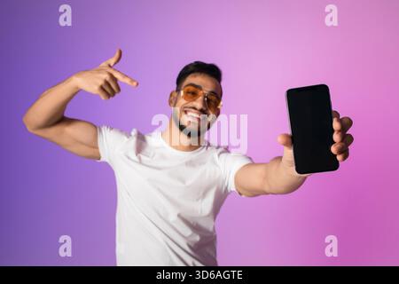 Young Arab Man Smiles and Points at Empty Smartphone Screen in Vibrant Neon Light Setting Stock Photo