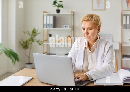 Mature doctor, focused attentive general practitioner in using laptop, giving online help, support Stock Photo