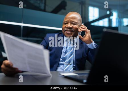 Mature black businessman reviewing document while talking on phone at his desk Stock Photo