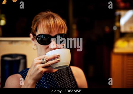 Woman wearing sunglasses drinking coffee at an outdoor café in Nice, France, captured in warm afternoon light. Stock Photo