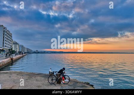 A fisherman greets the dawn on Thessaloniki’s quiet pier. Soft light rises over calm waters, as the city slowly stirs along the glowing waterfront. Stock Photo