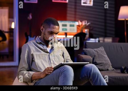 Focused african american guy multitasking on laptop in stylish living space, connected to the internet while managing his freelance work and remote responsibilities. Browsing web laying on sofa. Stock Photo