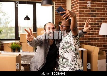 Excited multiethnic couple stand surrounded by moving boxes, taking break to capture selfie with cellphone. White bearded man makes peace sign as girlfriend holds mobile device to snap photo. Stock Photo