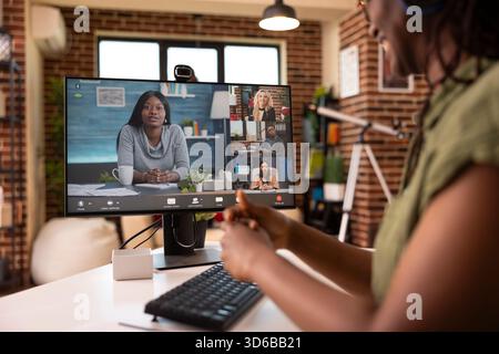 Cheerful black woman using digital device to manage team communication, happily discussing company profits. Female individual giving updates from home office during virtual meeting. Stock Photo